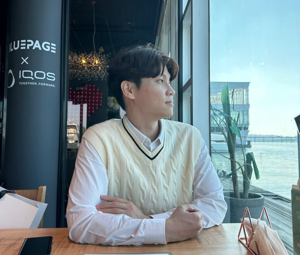 신비버스 창립자 '이서준'이 카페에 물이 보이는 카페에 앉아있다.날렵하지만 부드럽고 섬세한 외모를 가지고 있다.