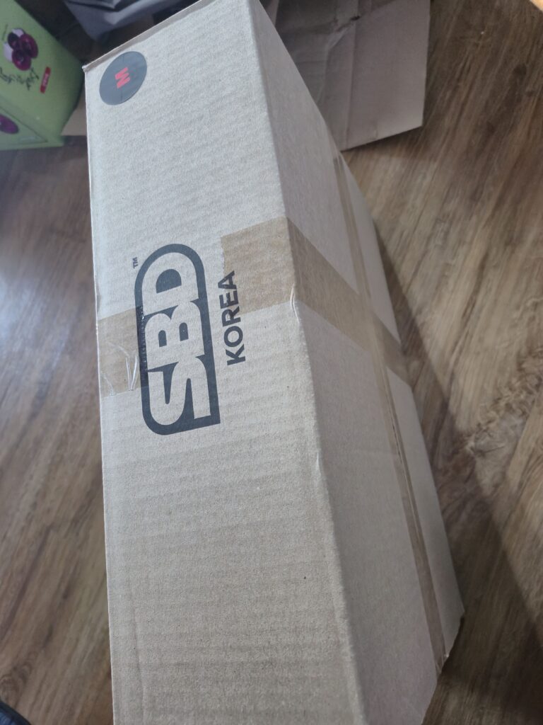 SBD KOREA 문구가 새겨진 네모난 박스. 한 쪽에는 'M'이라는 사이즈 표기 스티커가 붙어있다.